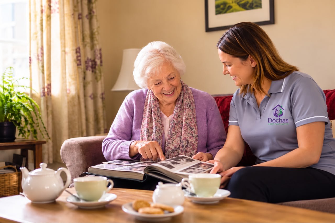 A Dòchas care worker sitting with a client on the sofa at home, smiling together as they look through a photo album, with tea laid out on the table